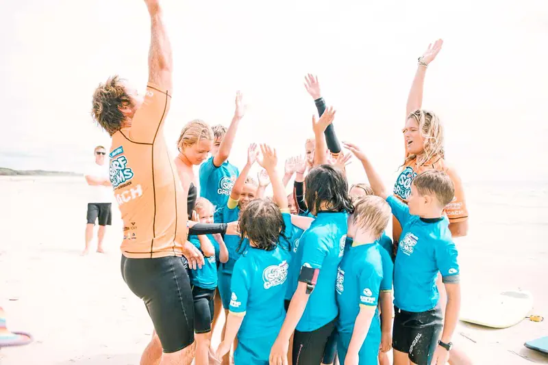 A coach high-fives a stoked group of young surfers in blue rash shirts