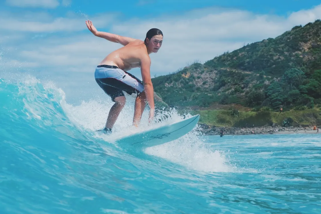 A teenage surfer trims across a clean turquoise wave beneath a bush-clad headland
