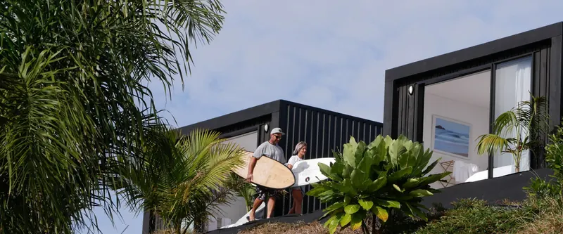 Guests carry surfboards past the Te Arai Eco Pods on the bush-lined path to the beach