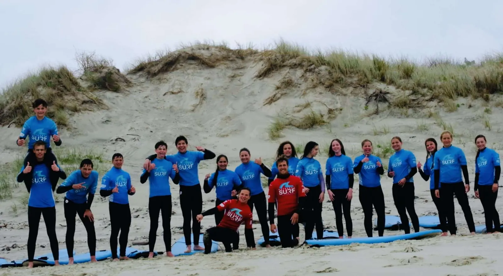 A full secondary group in blue rash shirts lines up on the sand at Te Arai