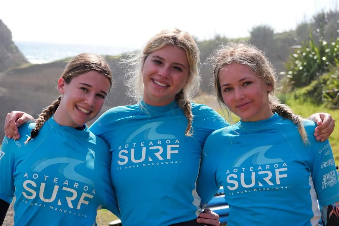 Three friends in Aotearoa Surf rash shirts arm in arm at the beach